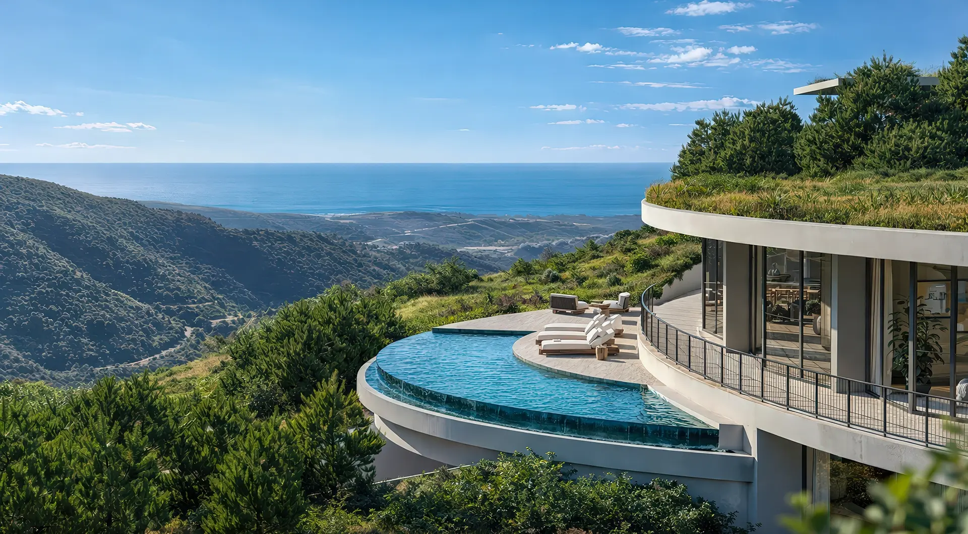 Contemporary circular villa with floor-to-ceiling glass walls and a green roof, perched on a hillside overlooking the ocean. A curved infinity pool extends from the terrace, surrounded by sun loungers and lush greenery with panoramic coastal views.