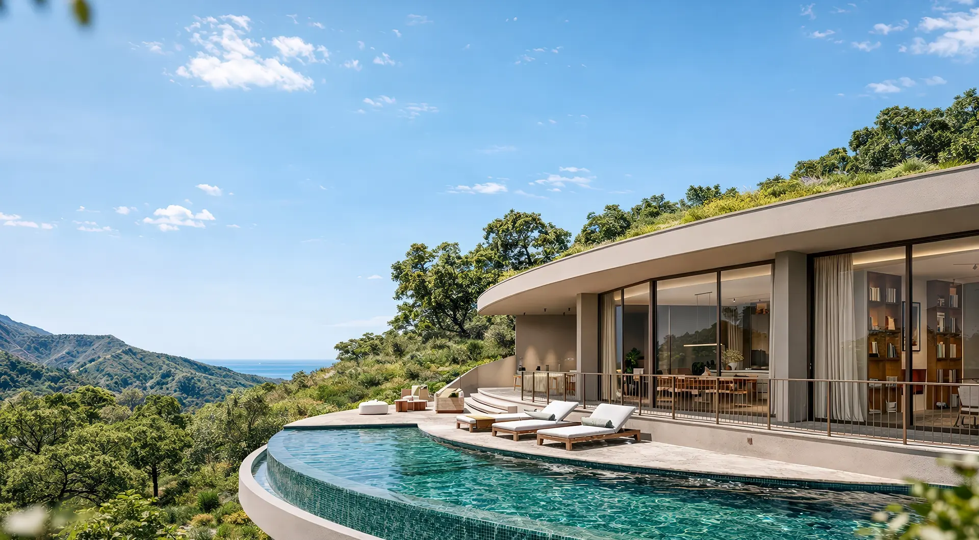 Modern curved villa with large floor-to-ceiling glass windows and a green roof, set on a hillside overlooking the ocean and rolling mountains. A sleek infinity pool wraps around the terrace, furnished with sun loungers and outdoor seating, surrounded by lush greenery under a clear blue sky.