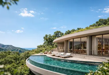 Modern curved villa with large floor-to-ceiling glass windows and a green roof, set on a hillside overlooking the ocean and rolling mountains. A sleek infinity pool wraps around the terrace, furnished with sun loungers and outdoor seating, surrounded by lush greenery under a clear blue sky.