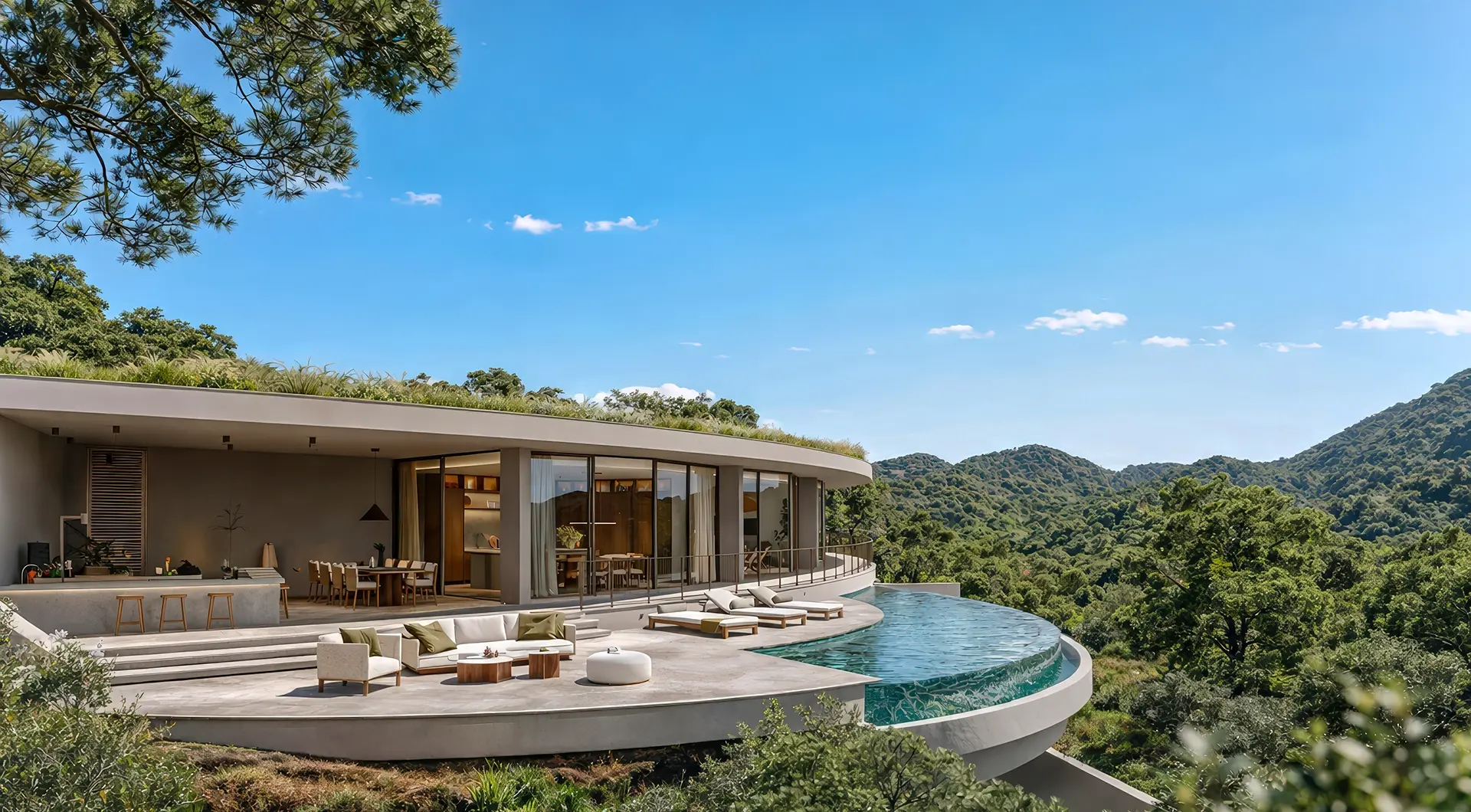 Contemporary single-story villa with curved architecture and floor-to-ceiling glass walls, set on a lush hillside. A spacious terrace with outdoor seating and dining area surrounds a curved infinity pool overlooking green mountains and dense forest under a clear blue sky.