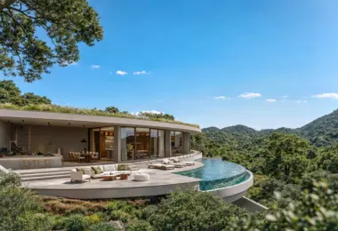 Contemporary single-story villa with curved architecture and floor-to-ceiling glass walls, set on a lush hillside. A spacious terrace with outdoor seating and dining area surrounds a curved infinity pool overlooking green mountains and dense forest under a clear blue sky.
