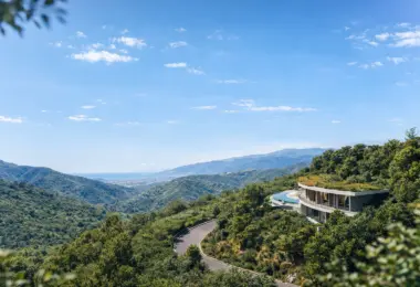 Contemporary curved villa with a green roof and expansive glass walls, nestled on a lush mountainside. The home overlooks rolling green hills, a winding road, and distant ocean views under a clear blue sky, creating a secluded luxury retreat in nature.