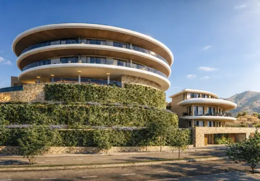 Striking modern residential development in Fuengirola Carvajal featuring curved multi-level architecture, expansive glass-fronted terraces, natural stone façades, lush vertical greenery, and scenic mountain surroundings under a clear Mediterranean sky.