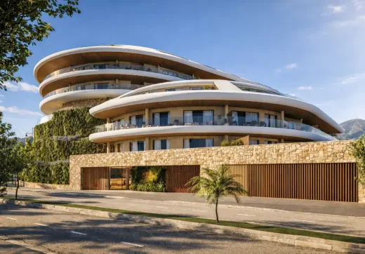 Distinctive contemporary residential complex in Fuengirola Carvajal featuring flowing curved architecture, expansive glass terraces, natural stone façades, lush vertical greenery, and panoramic Mediterranean surroundings under a clear blue sky.